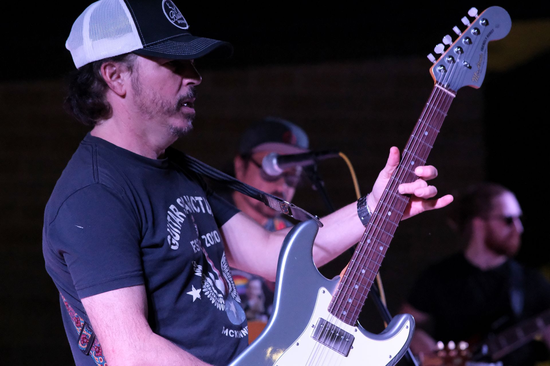 Man playing electric guitar onstage, wearing a hat and glasses, with bandmates blurred in the background.
