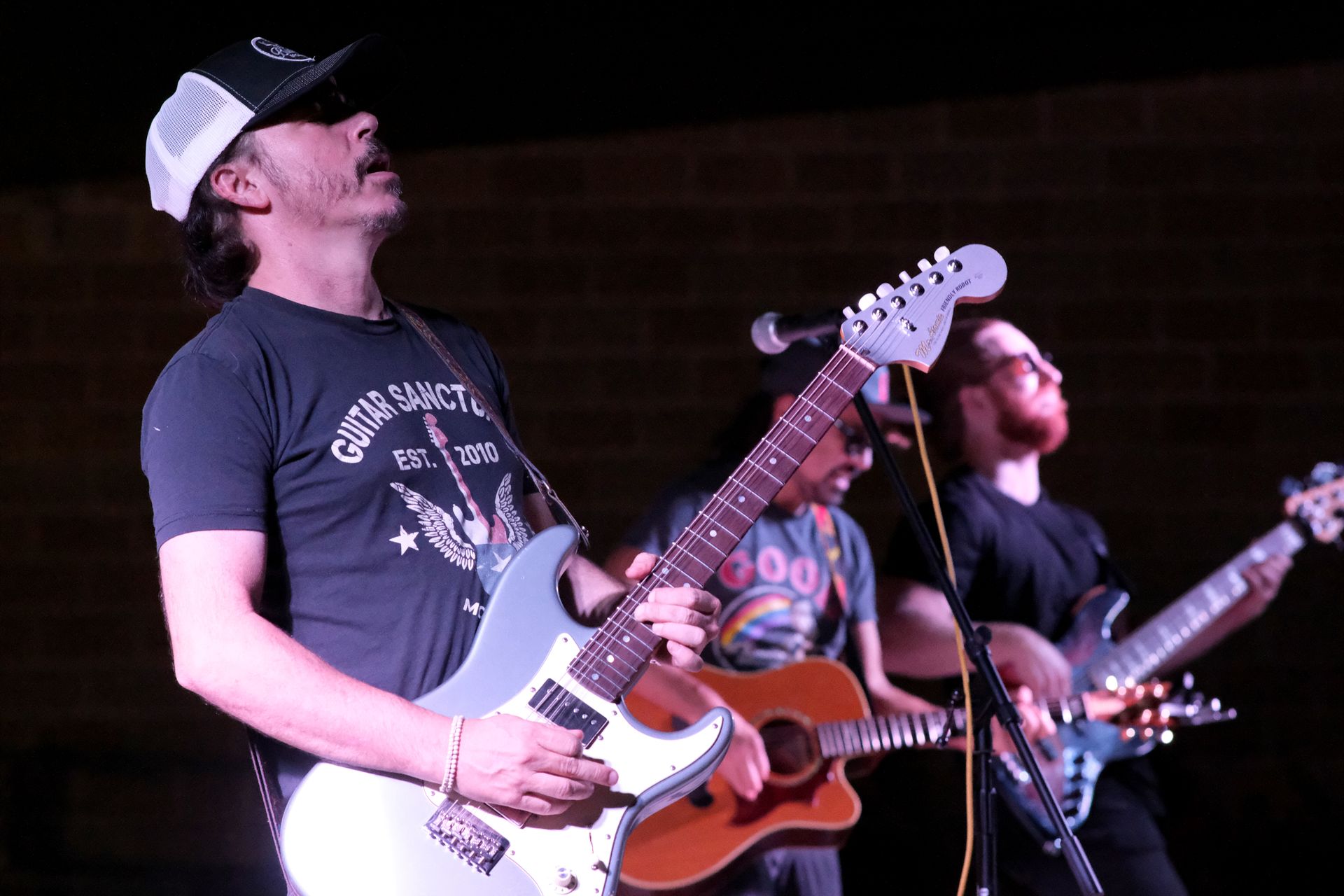 Band performing on stage, guitarist playing a light blue electric guitar, other musicians visible.
