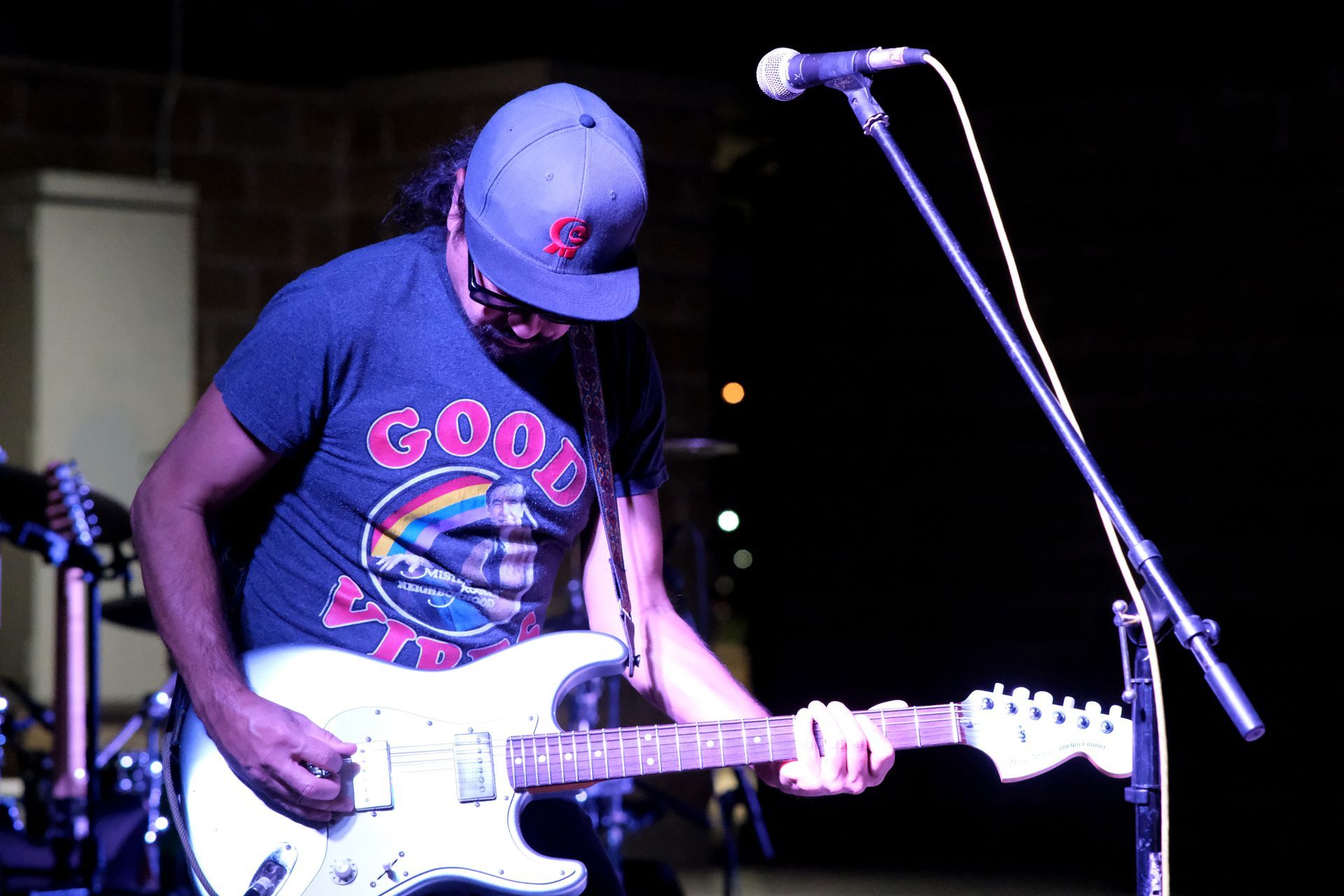 Guitarist playing on stage, wearing a blue cap and graphic t-shirt, with a microphone in front of him.