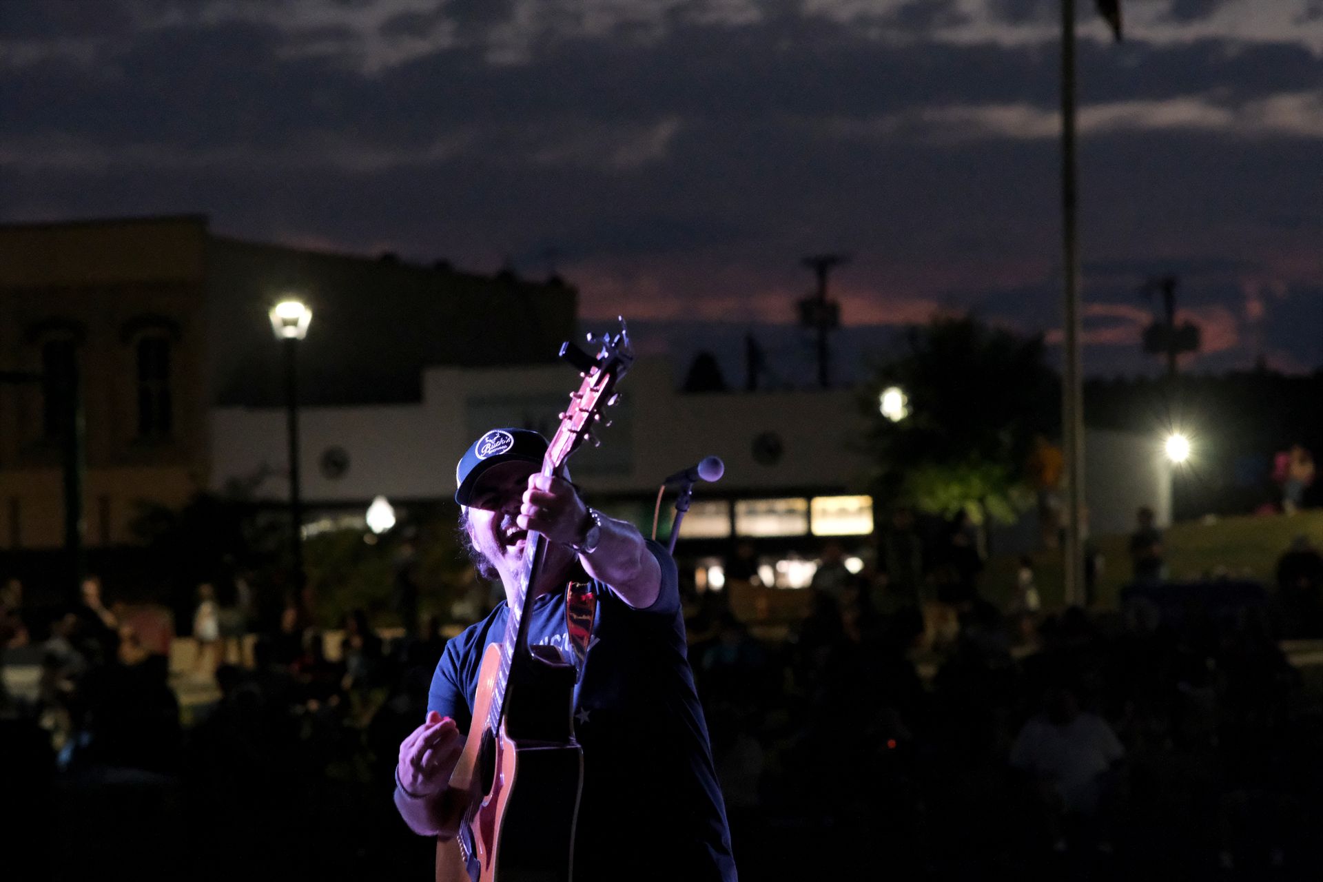 Musician on stage at night, playing guitar, with a crowd in the background. Buildings and streetlights visible.
