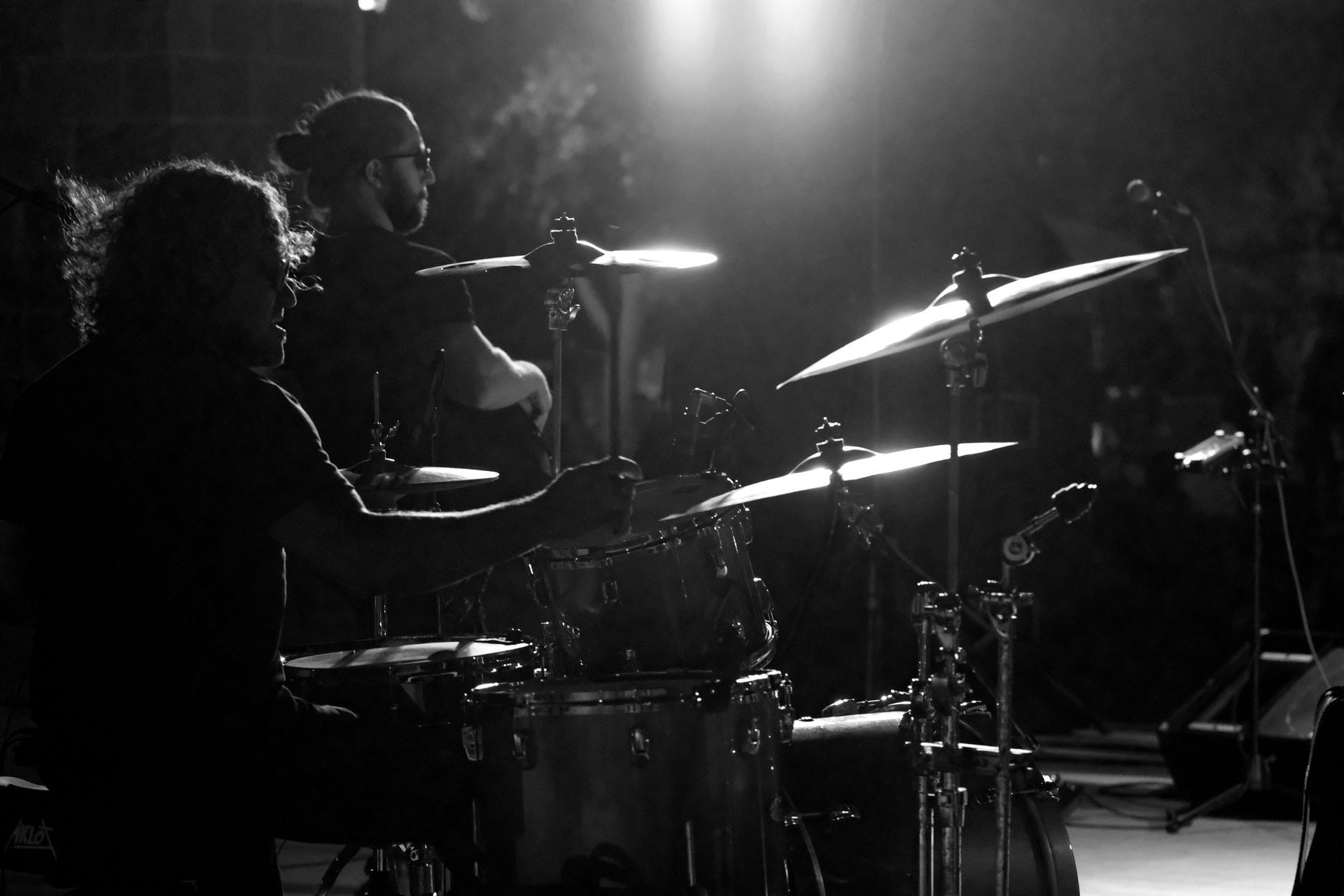Drummer performing onstage, silhouetted in black and white, stage lights illuminate cymbals.