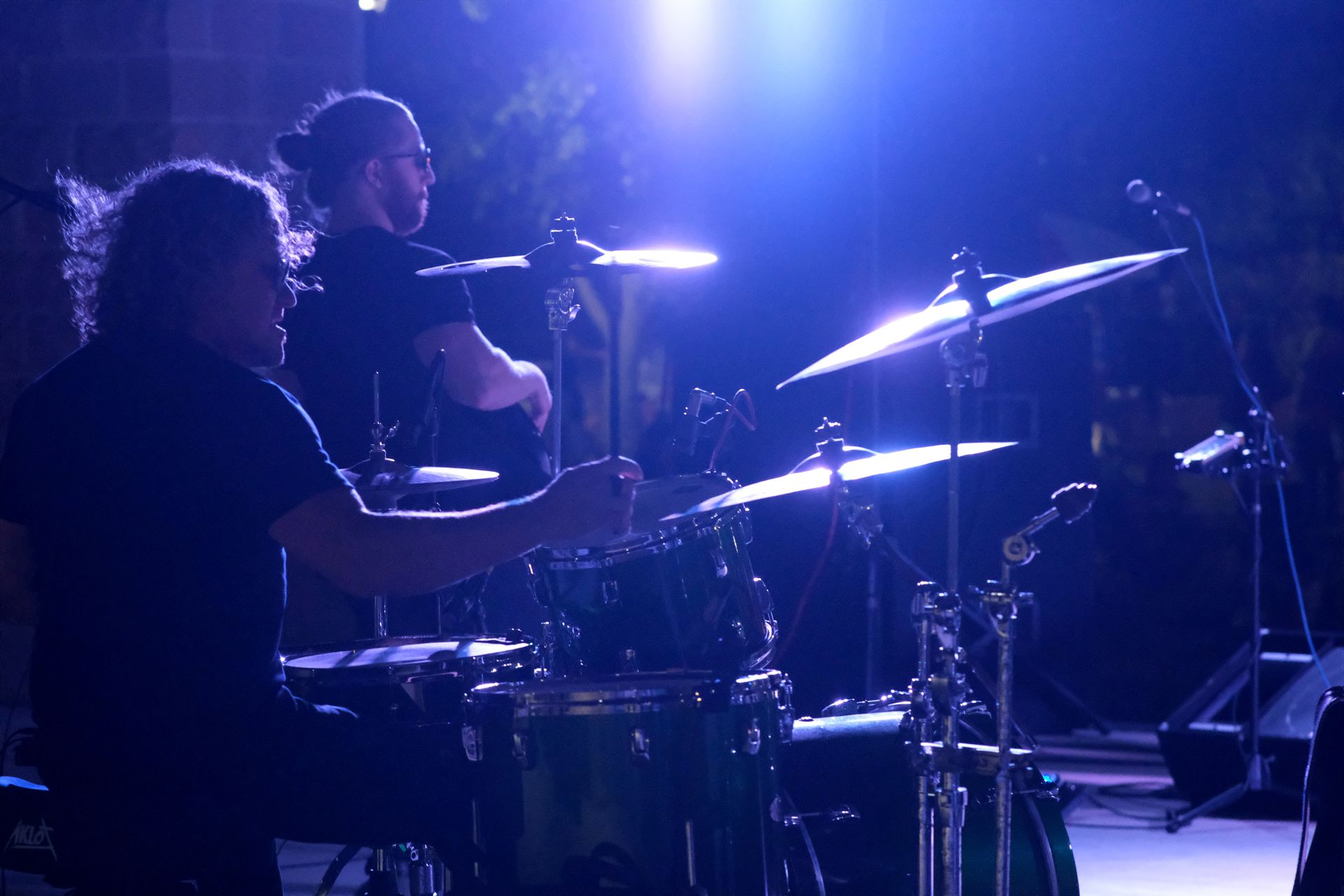 Drummer performing on stage under blue stage lights; another musician stands behind.