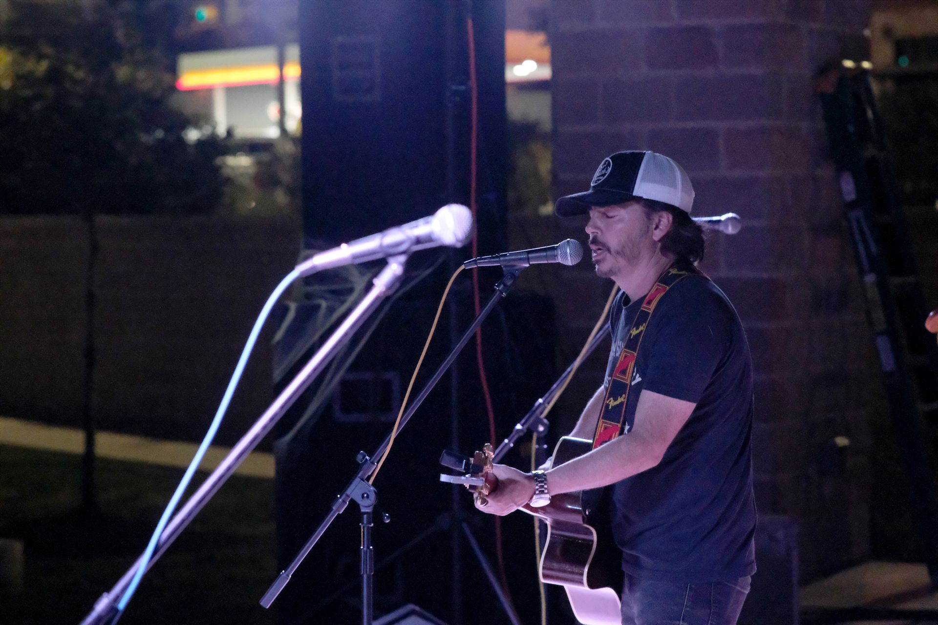 Man with guitar singing at an outdoor concert, wearing a hat, black shirt, and jeans, with microphones.