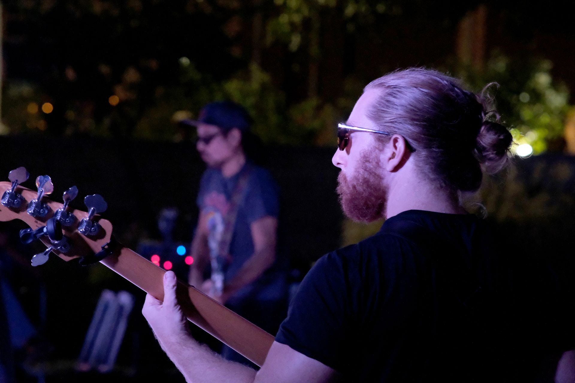 Man with beard, sunglasses, and ponytail playing bass guitar at outdoor night performance. Another musician in background.