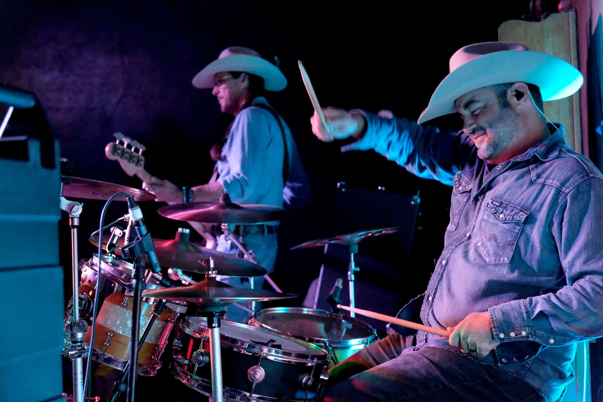 Two musicians in cowboy hats perform on a stage; one plays drums, the other bass.