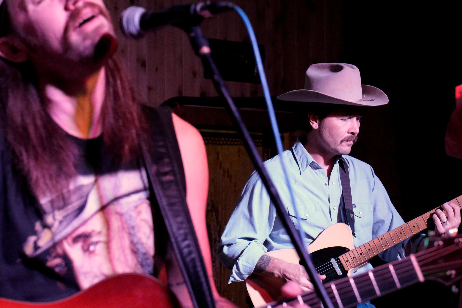 Two guitarists performing on stage; one in a cowboy hat, the other wearing a graphic t-shirt.