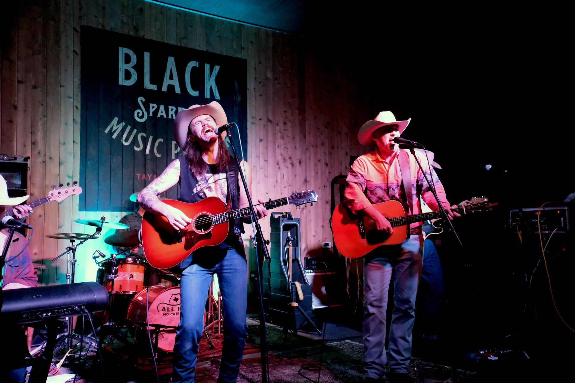 Band performing onstage at Black Sparrow Music Parlor; two guitarists singing with cowboy hats, drums in the background.