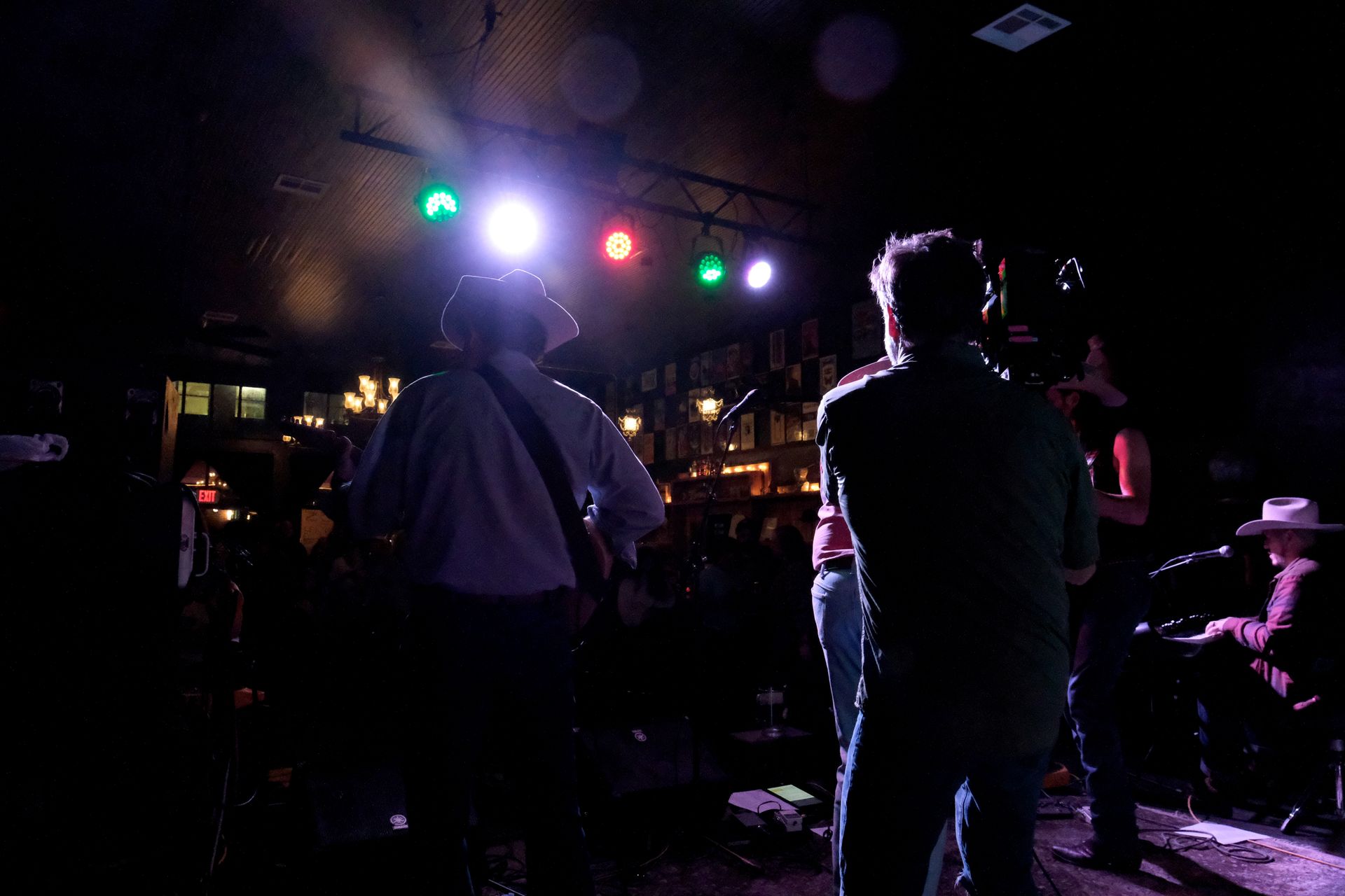 Band performing on stage in a dark bar, bathed in colored stage lights. Audience silhouettes in foreground.