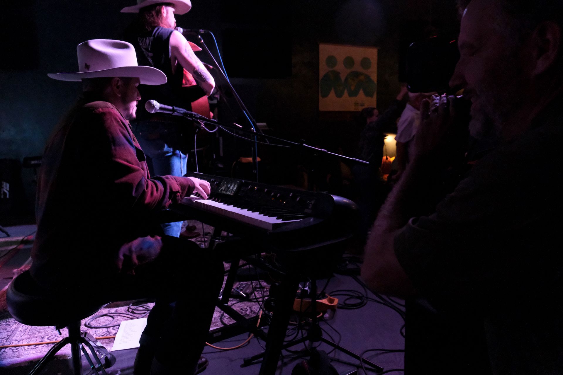 Musician playing a keyboard on stage, wearing a cowboy hat. Another musician stands nearby, with an audience member close by.