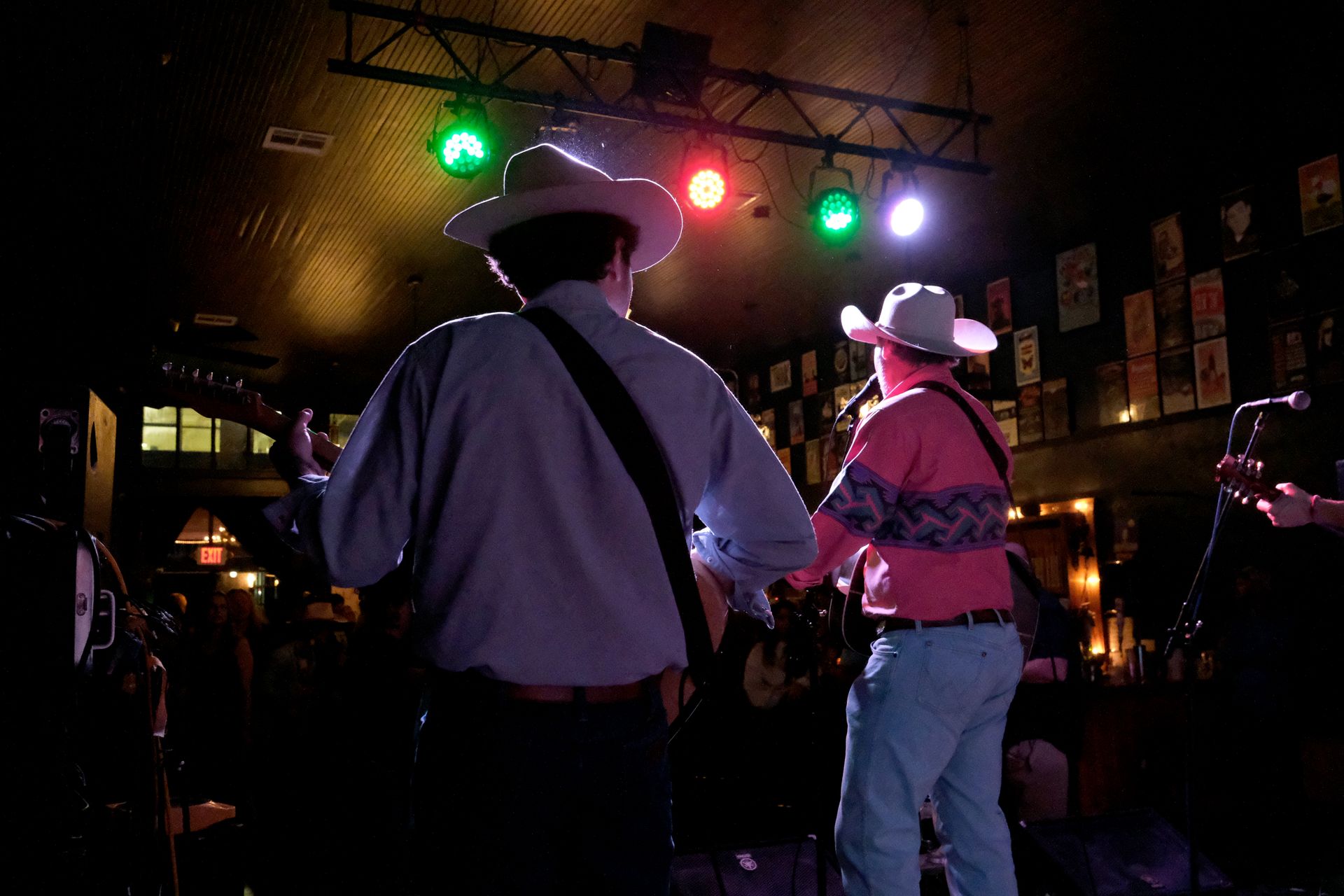 Two guitarists wearing cowboy hats performing on a stage lit by colorful lights in a dimly lit venue.