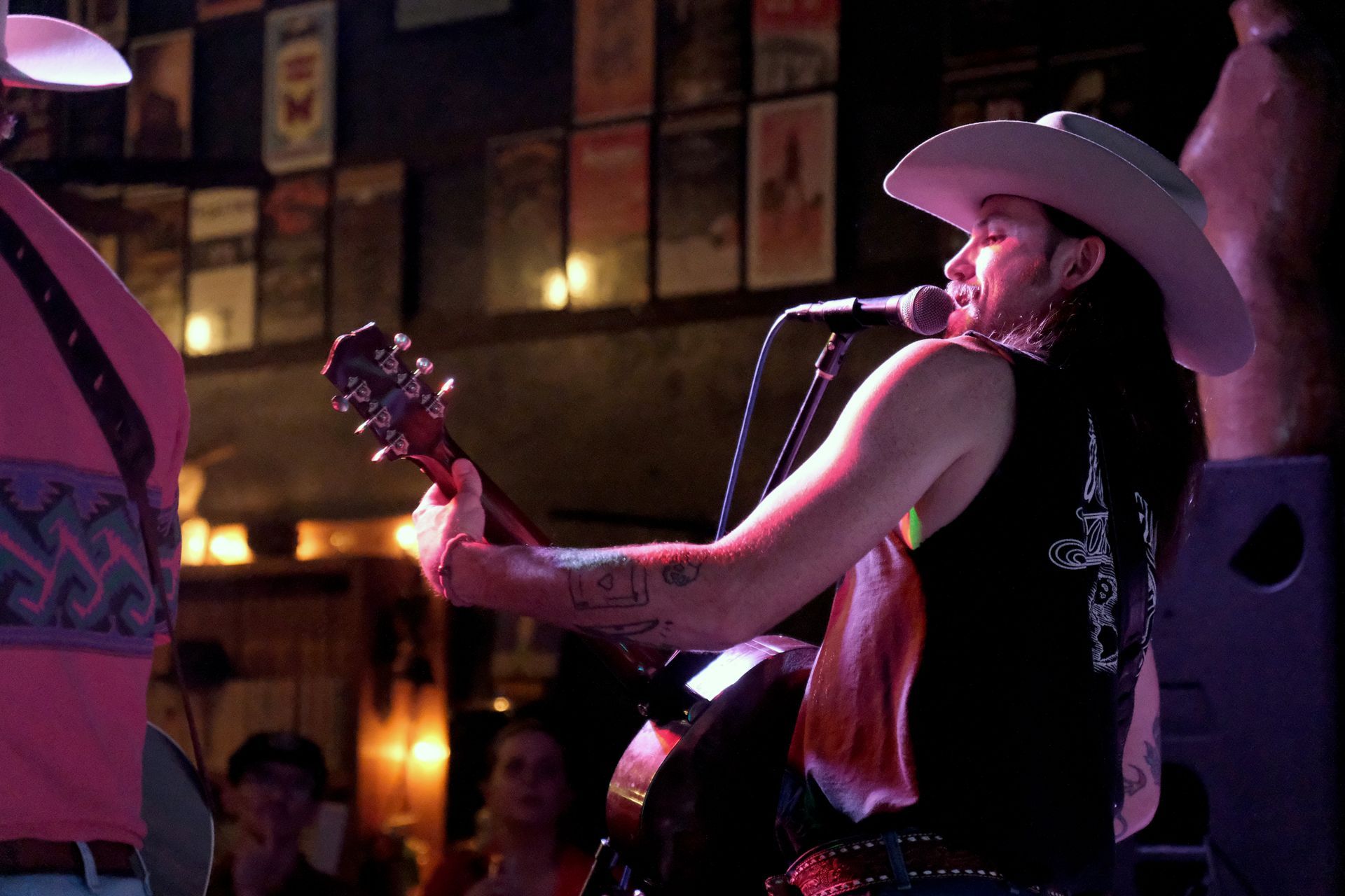 Man in cowboy hat plays guitar on stage, singing into a microphone. Dark interior with posters.