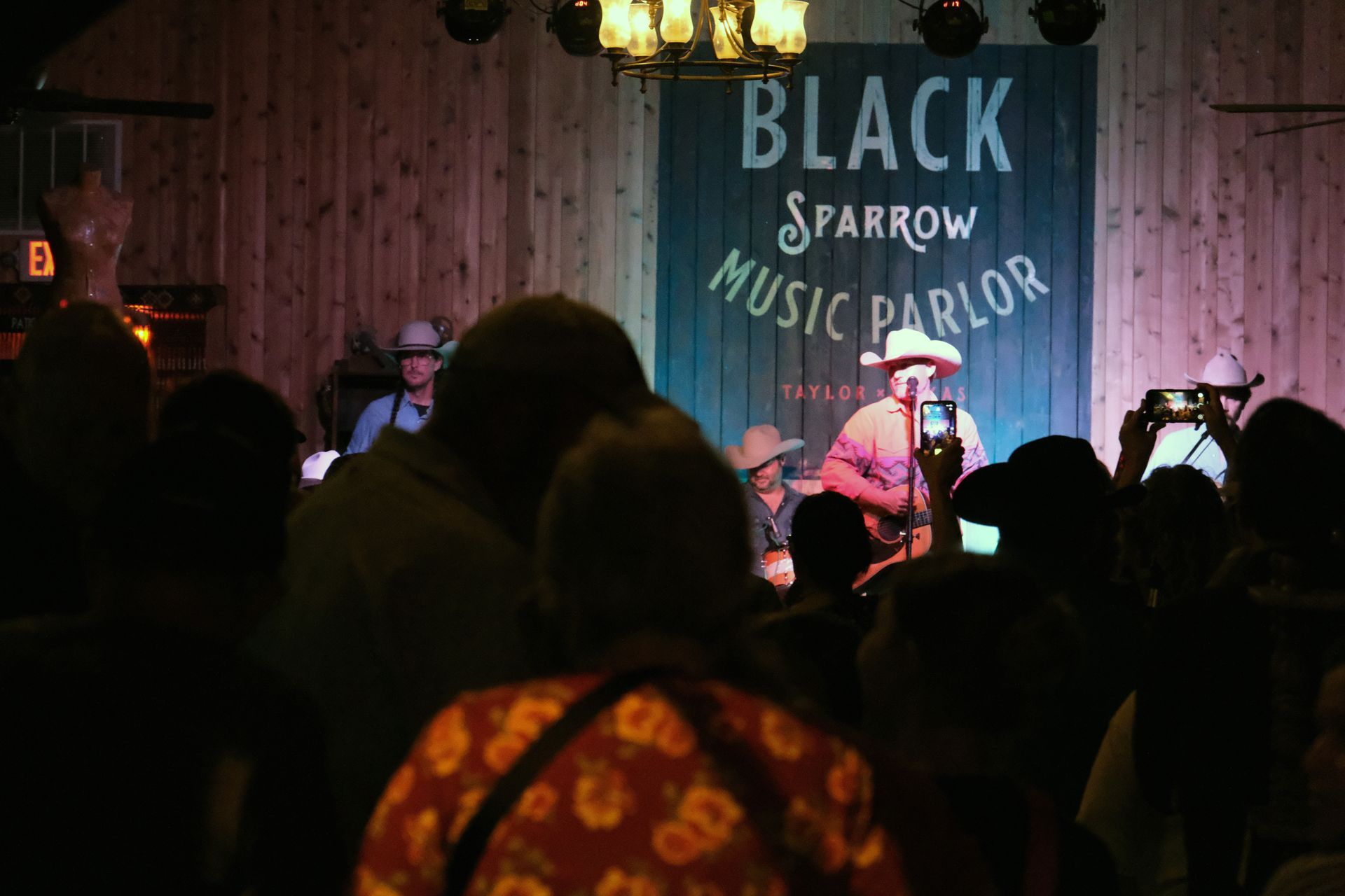 Band performing at Black Sparrow Music Parlor. Audience members watch, some taking photos. Dark setting, stage lit.