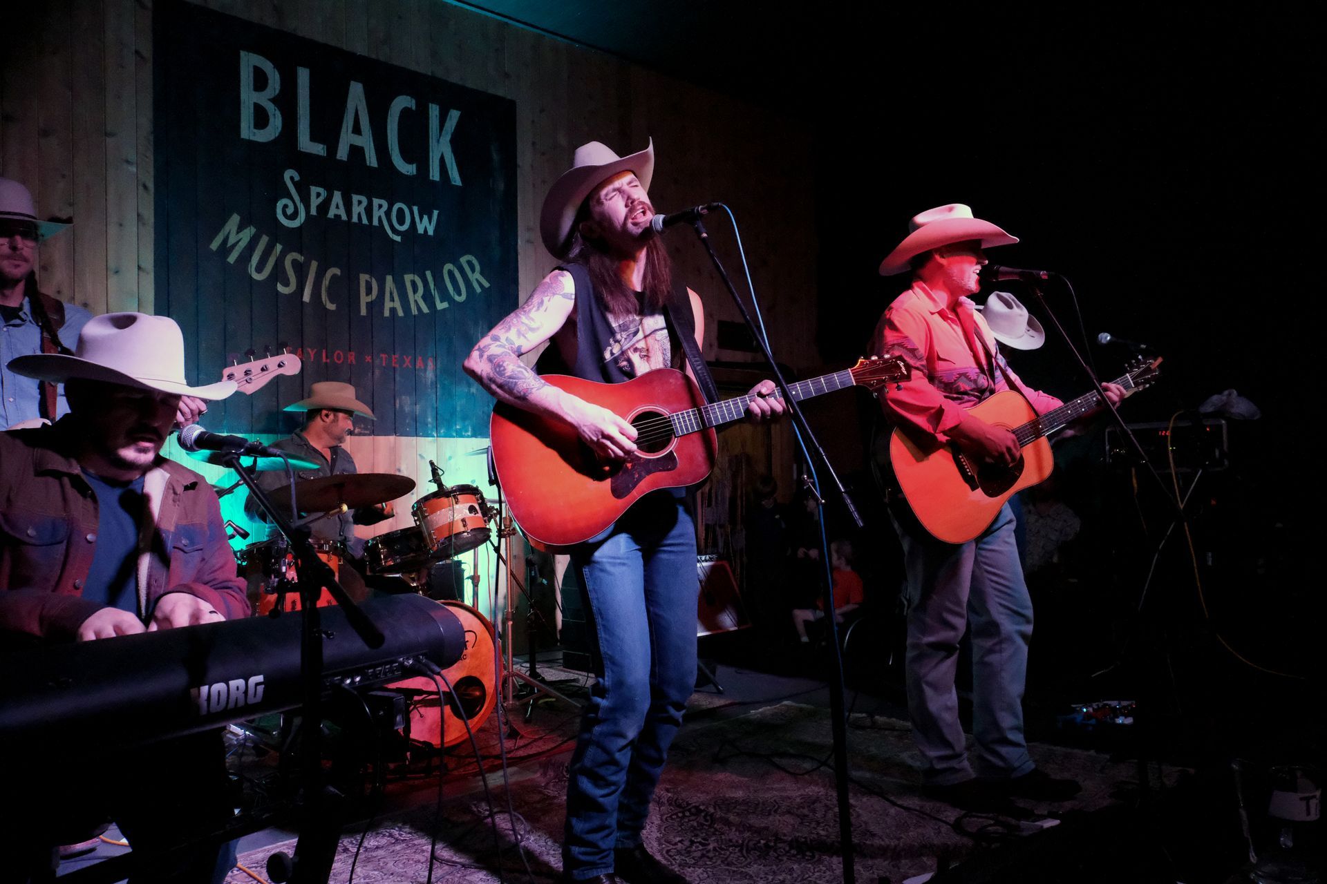 A band performing at the Black Sparrow Music Parlor. Musicians play guitars, keyboard, and drums, wearing cowboy hats.