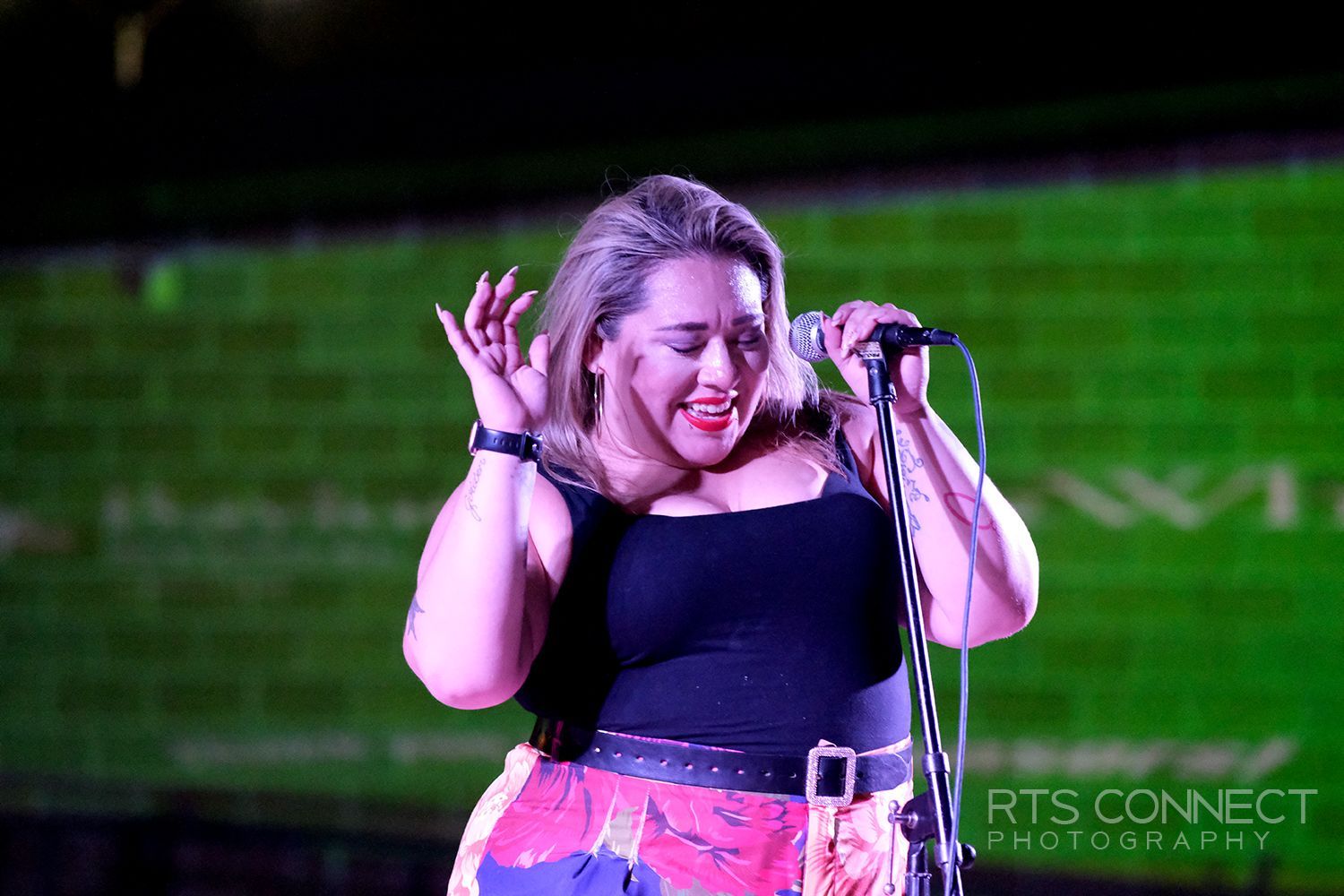 Woman singing into a microphone onstage; wearing black top, patterned skirt, brick wall backdrop.