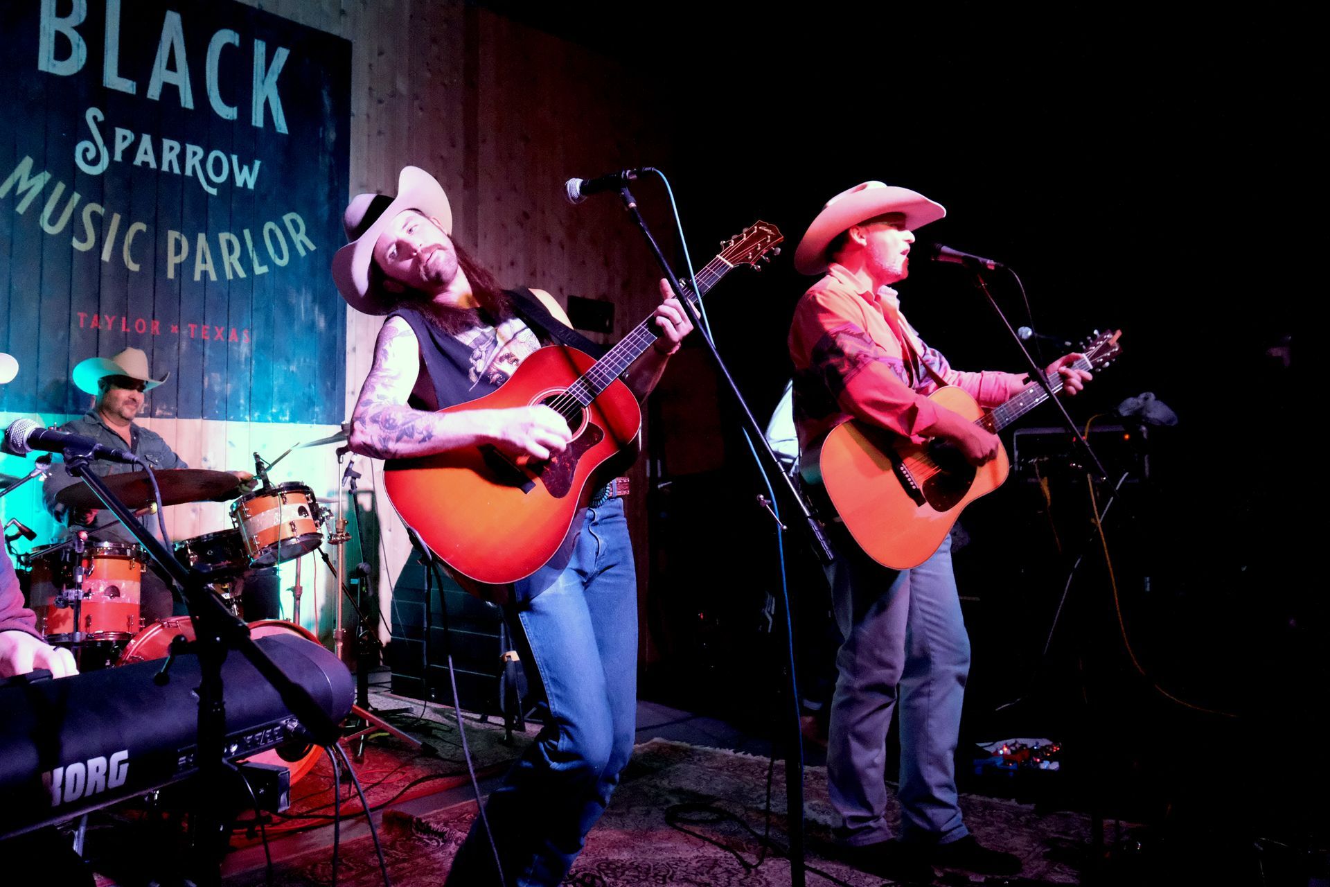 Band performing on stage at Black Sparrow Music Parlor; two guitarists singing, drummer visible in the background.