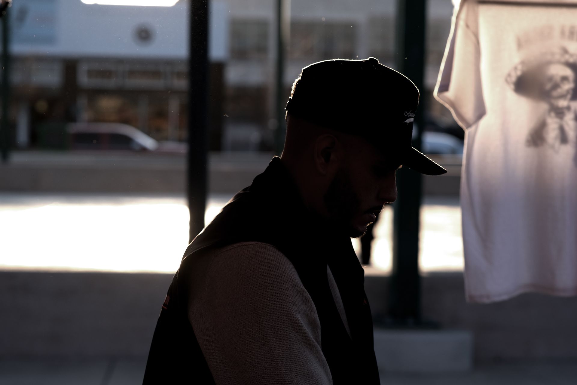 Man in cap and vest, looking down, near a window with a white t-shirt hanging.