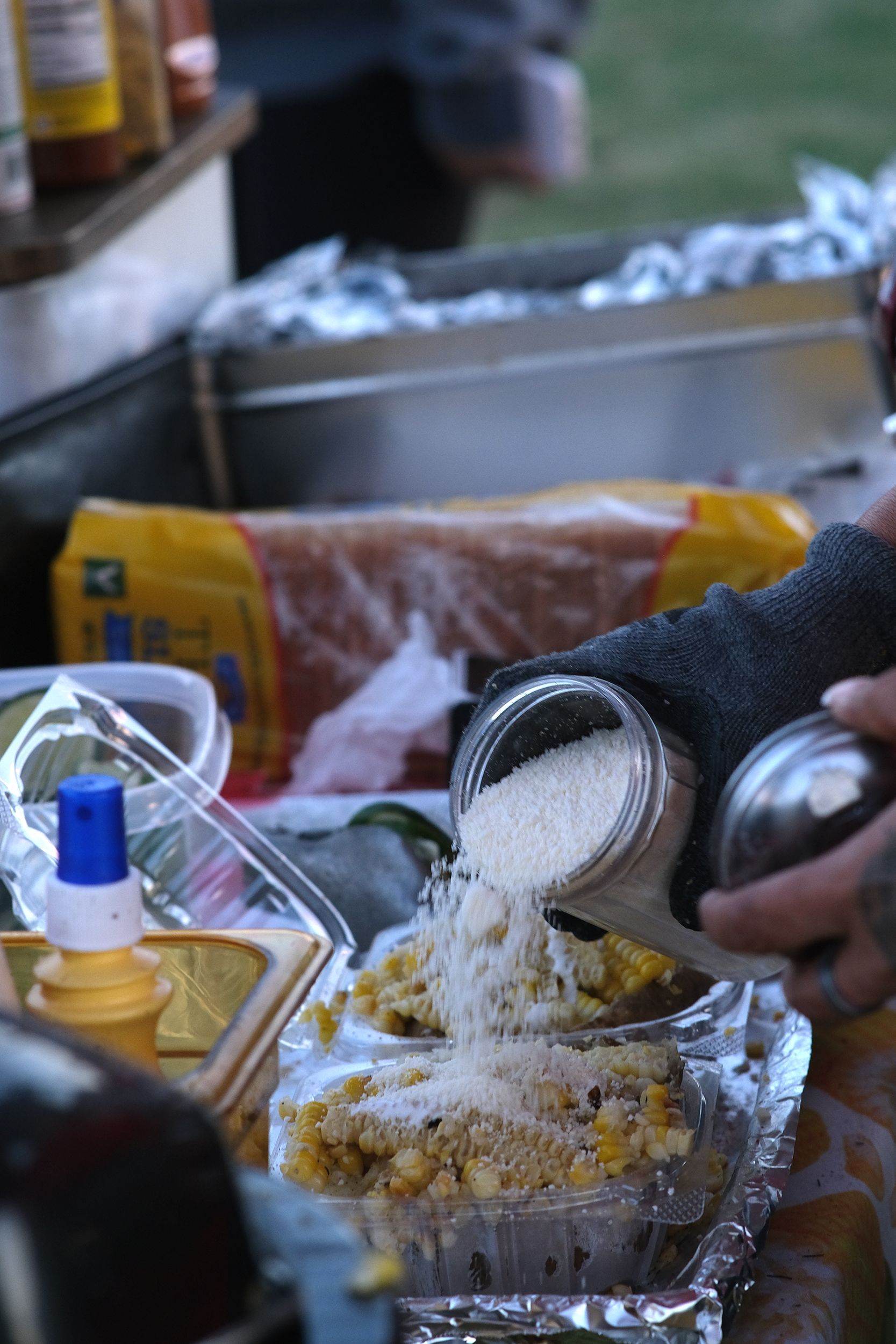 Person sprinkles grated cheese on corn in a container, outdoors.