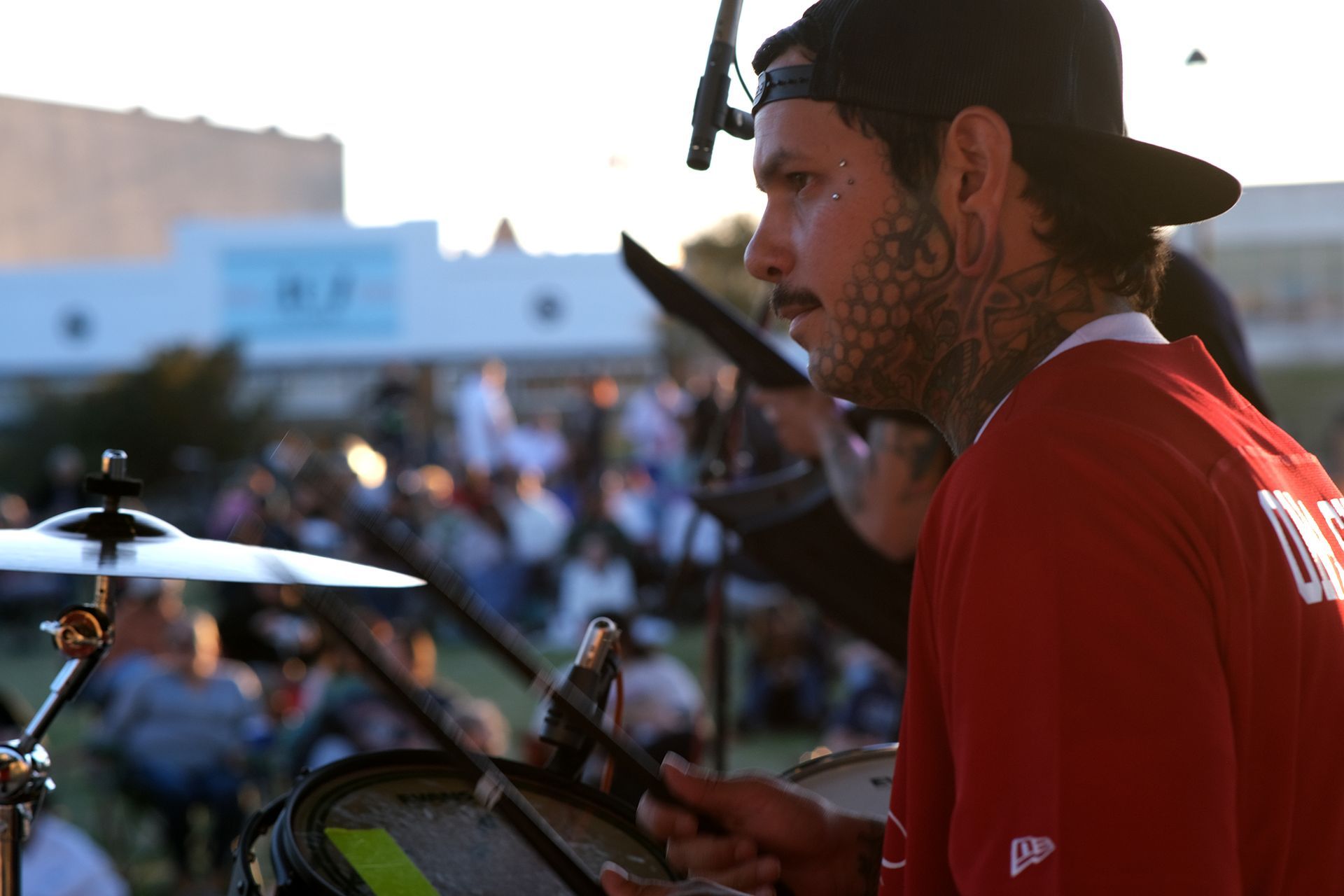 Drummer playing outdoors, wearing a red shirt and black cap. Crowd in background.