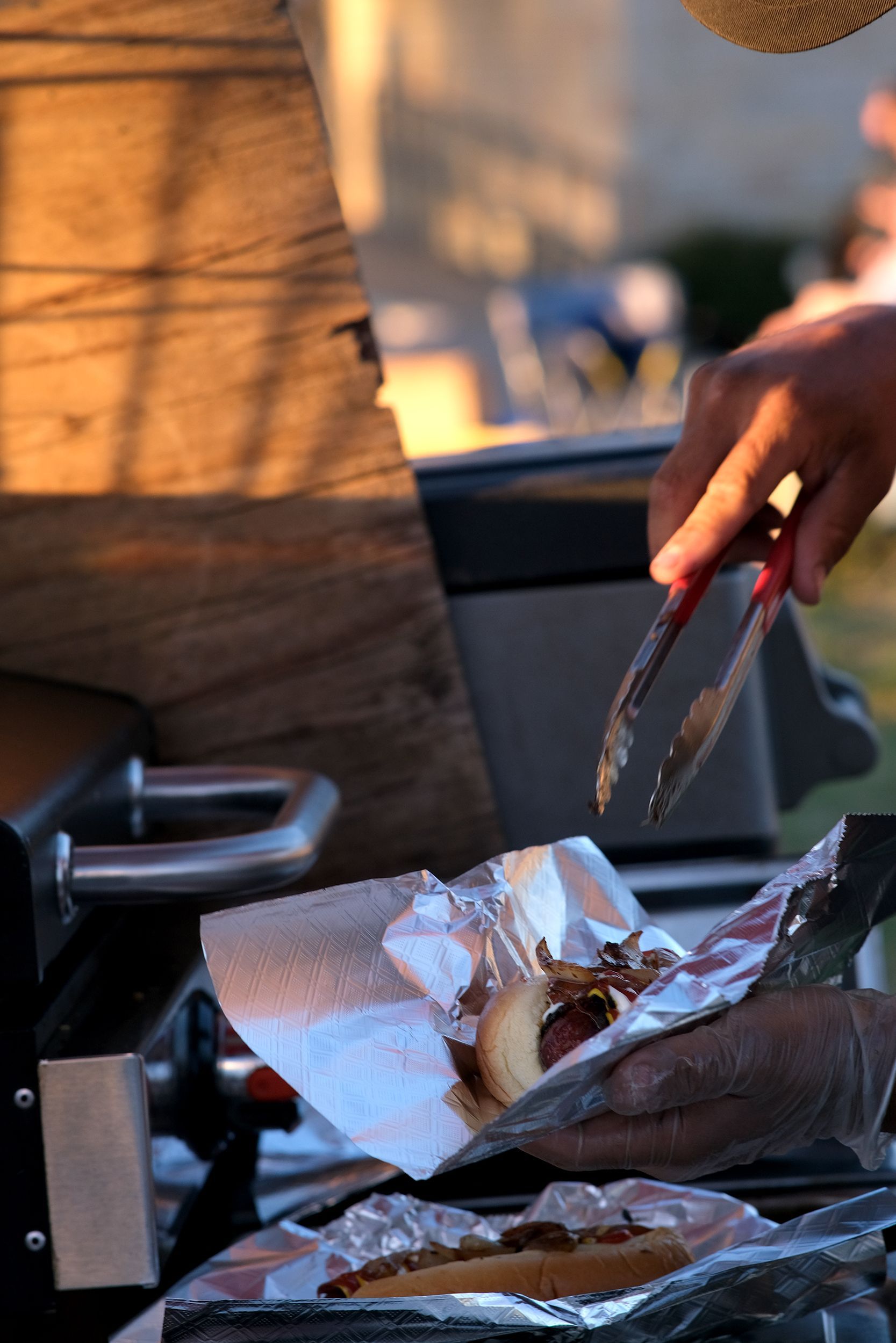 Person using tongs to place a cooked hotdog with onions into foil wrapping on a grill.