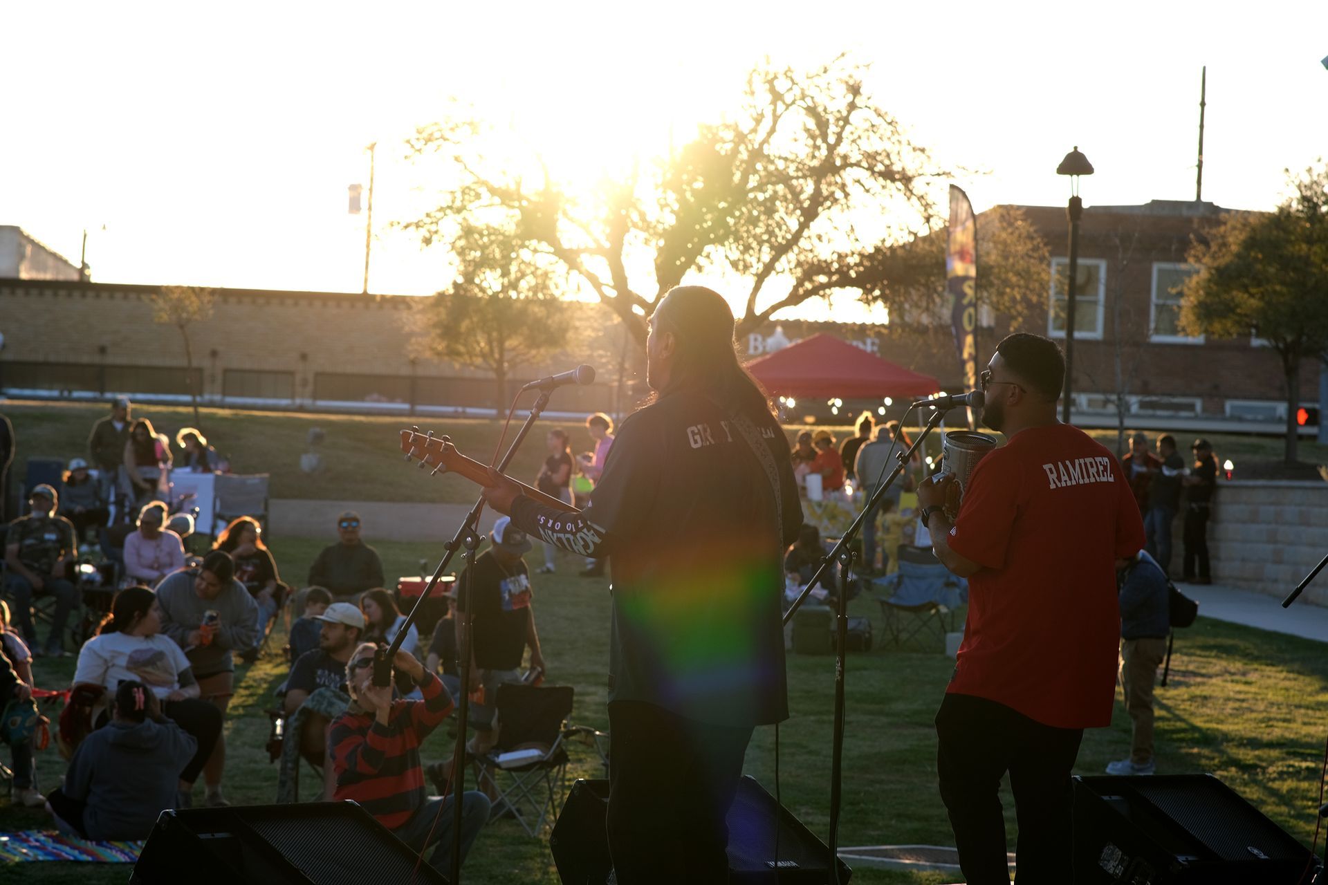 A band performs outdoors at sunset. A crowd sits on the grass, watching.