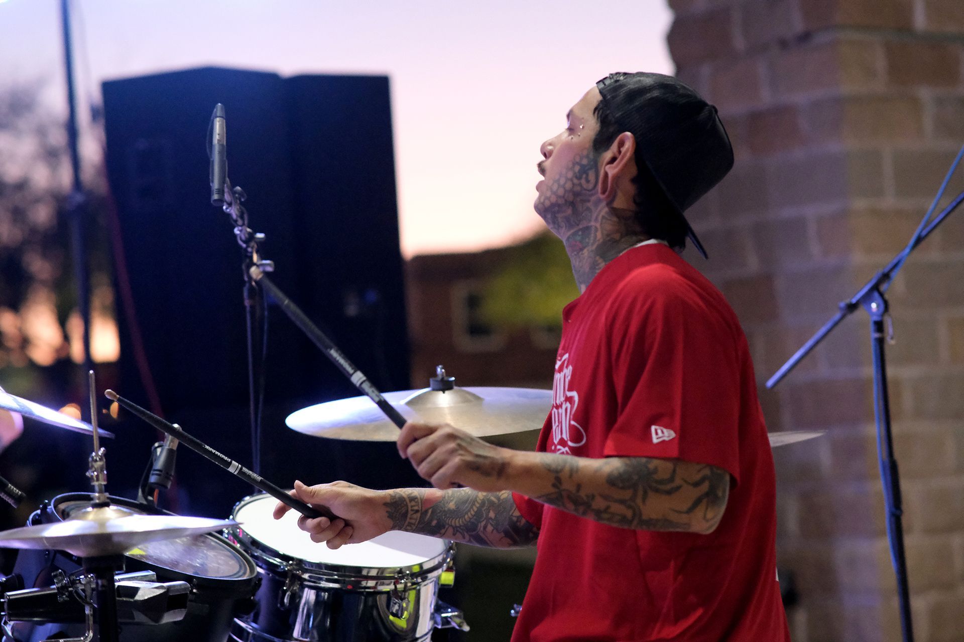 Drummer playing drums outside at dusk, wearing a red shirt and a black hat.