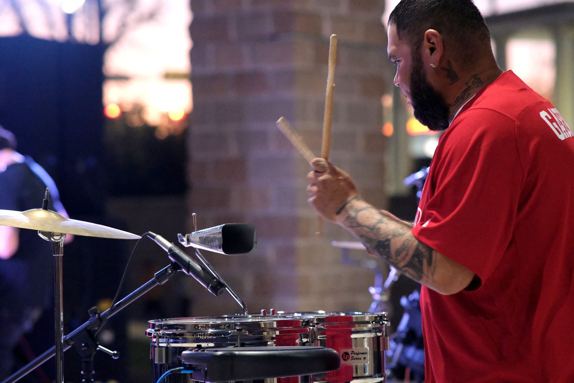 Man in red shirt playing a drum set outdoors with drumsticks.
