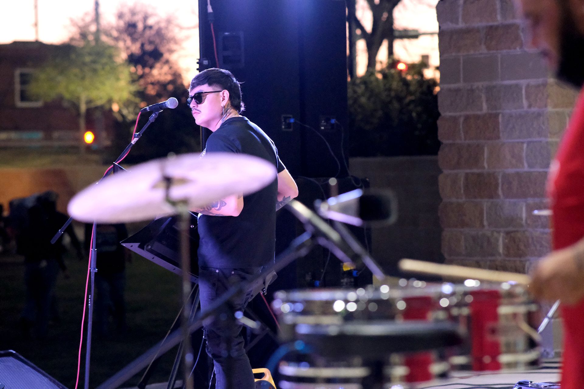 A person singing with a microphone at an outdoor concert, with drums and sound equipment.