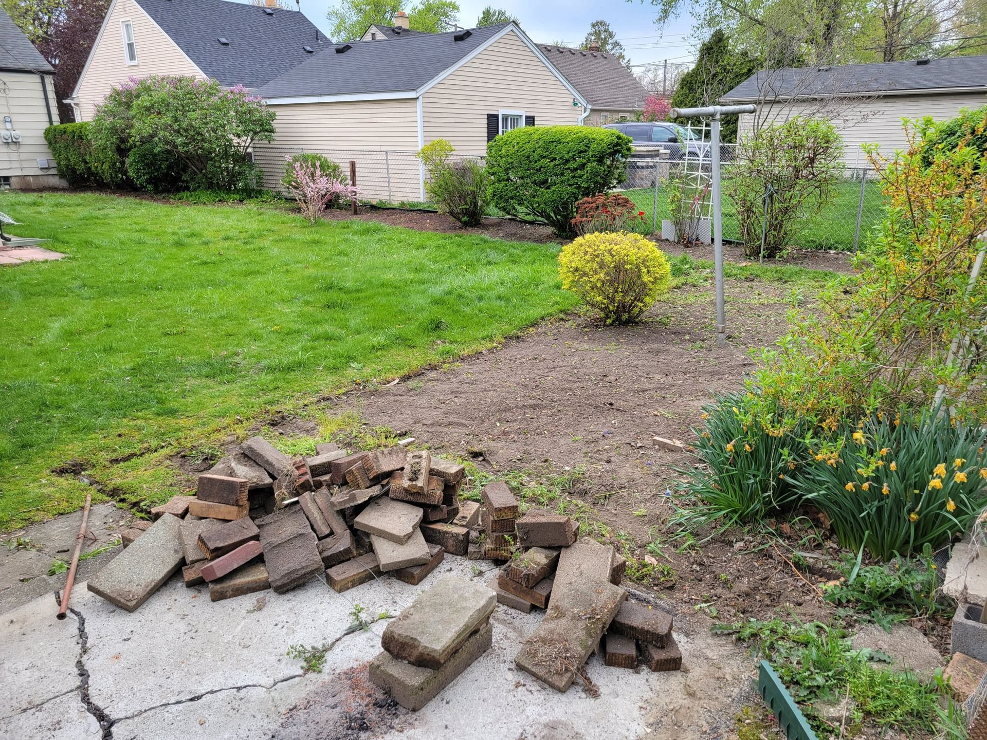 Backyard scene with grass, garden bed, and pile of bricks.