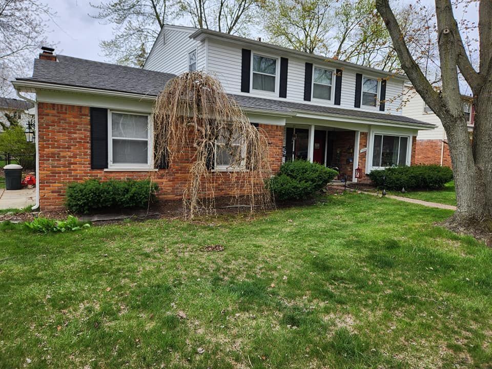 Two-story brick and white house with black shutters, green lawn, and dead weeping tree in front yard.