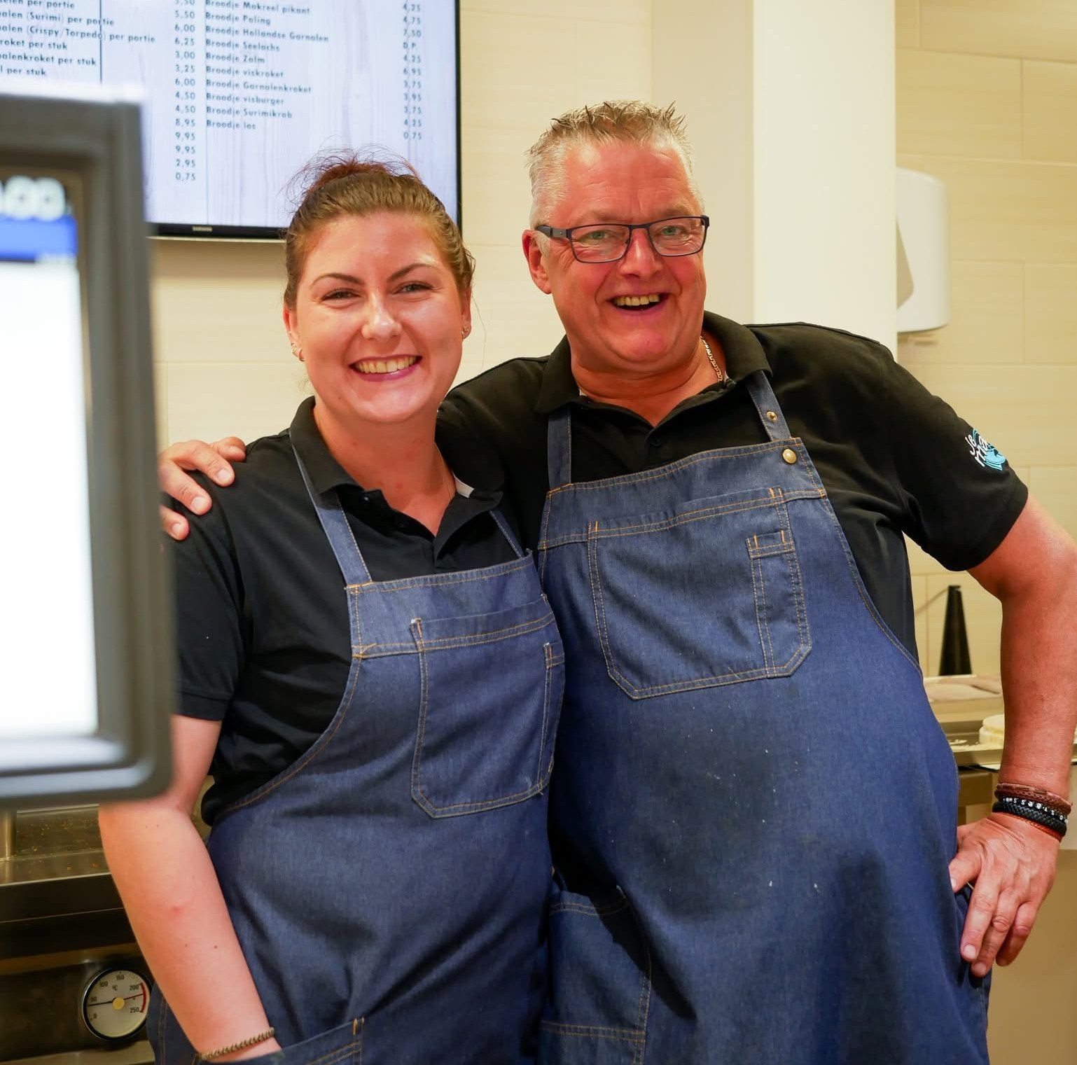 Twee mensen in zwarte shirts en glimlachen naar elkaar in een horecagelegenheid, met een menubord op de achtergrond.