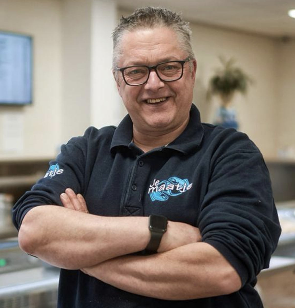 Een persoon met een bril en een donker poloshirt met logo staat met de armen over elkaar in een binnenruimte.