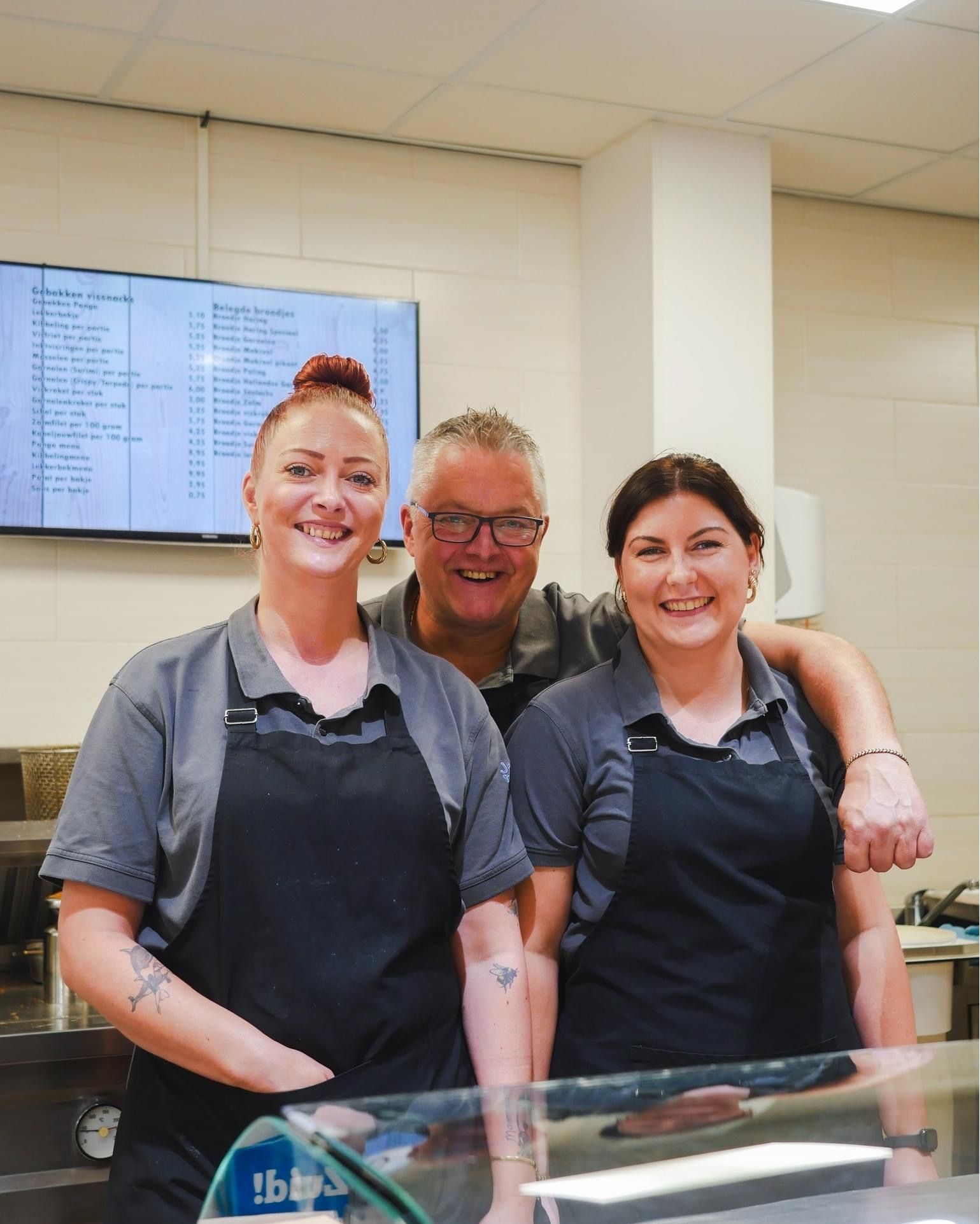 Drie personeelsleden in grijze shirts en zwarte schorten glimlachen achter een servicebalie in een horecagelegenheid.