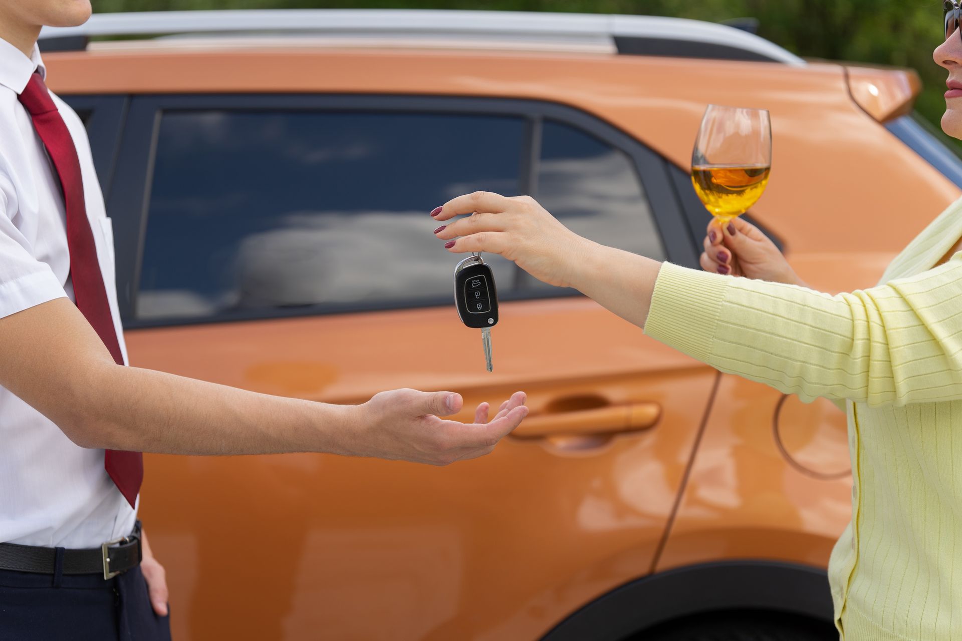 A person offers car keys to another beside an orange SUV, with a glass of orange drink in the foreground