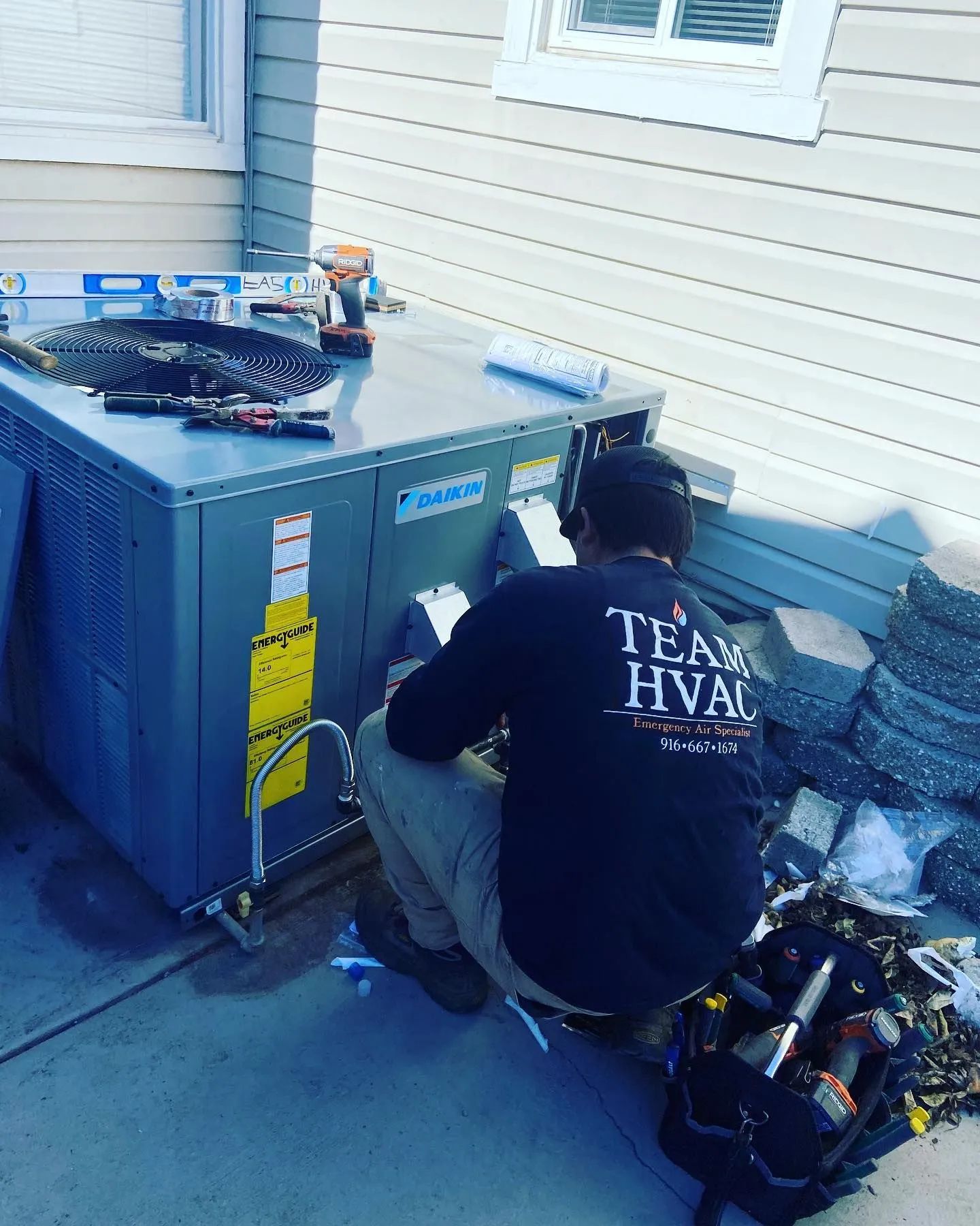 Technician working on a blue HVAC unit beside a house, with tools and equipment nearby.