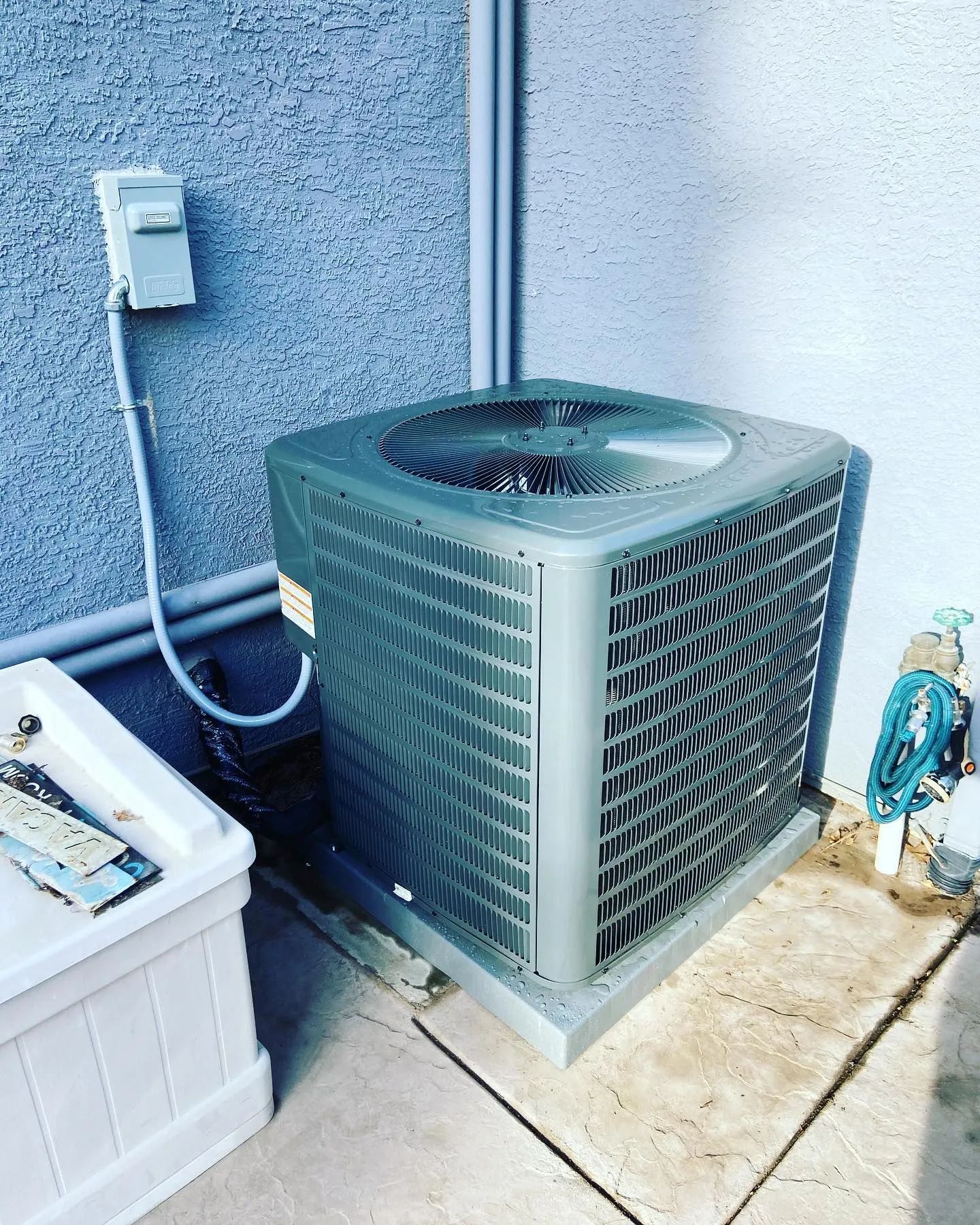 Air conditioning unit next to a white box and blue wall. Gray unit with fan.