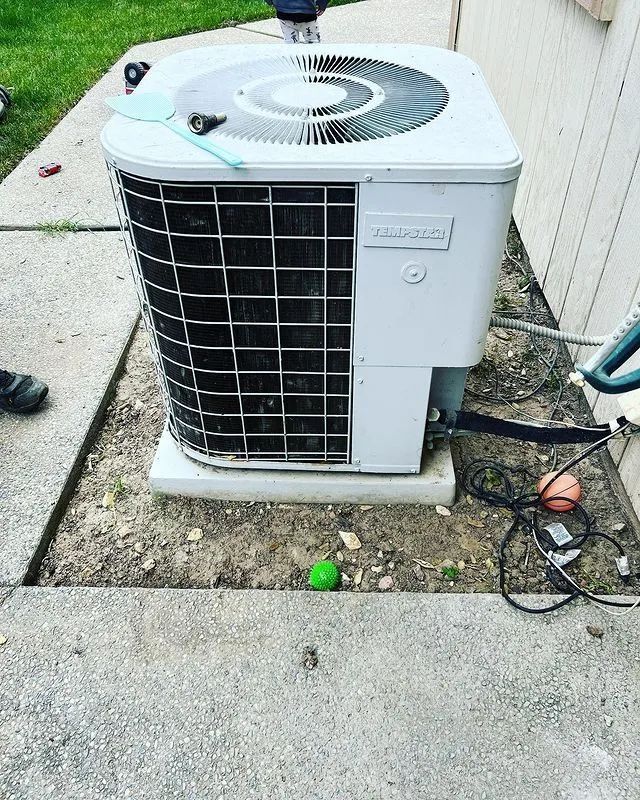 An air conditioning unit on a concrete pad next to a wall. Various tools are nearby.