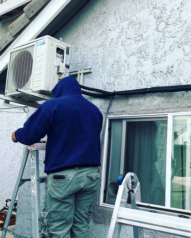 A man is standing on a ladder installing an air conditioner on the side of a building.