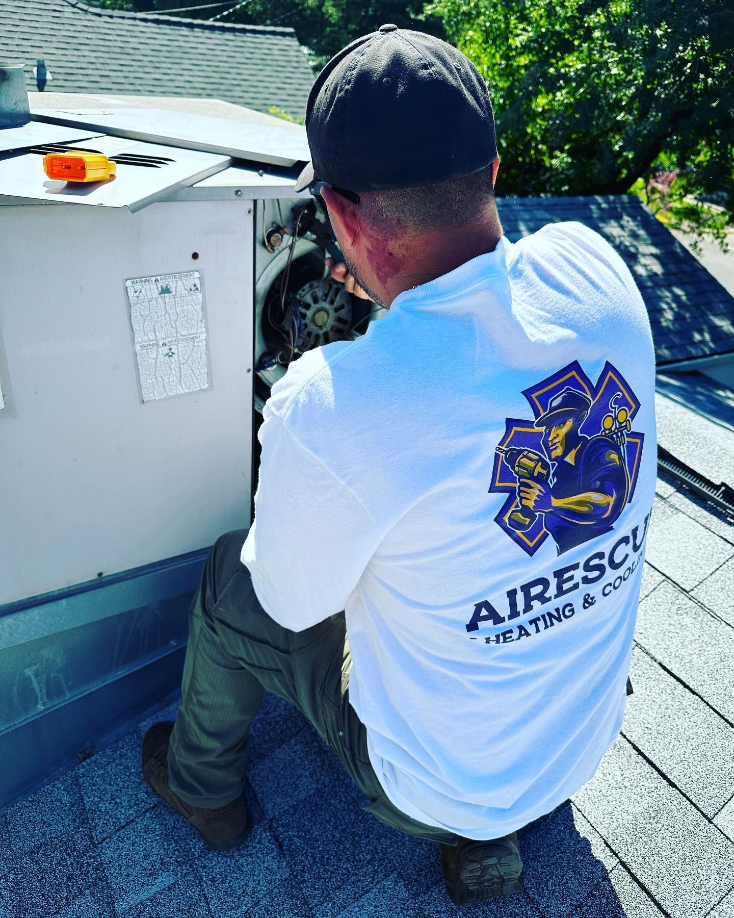 A man is kneeling on a roof working on an air conditioner.
