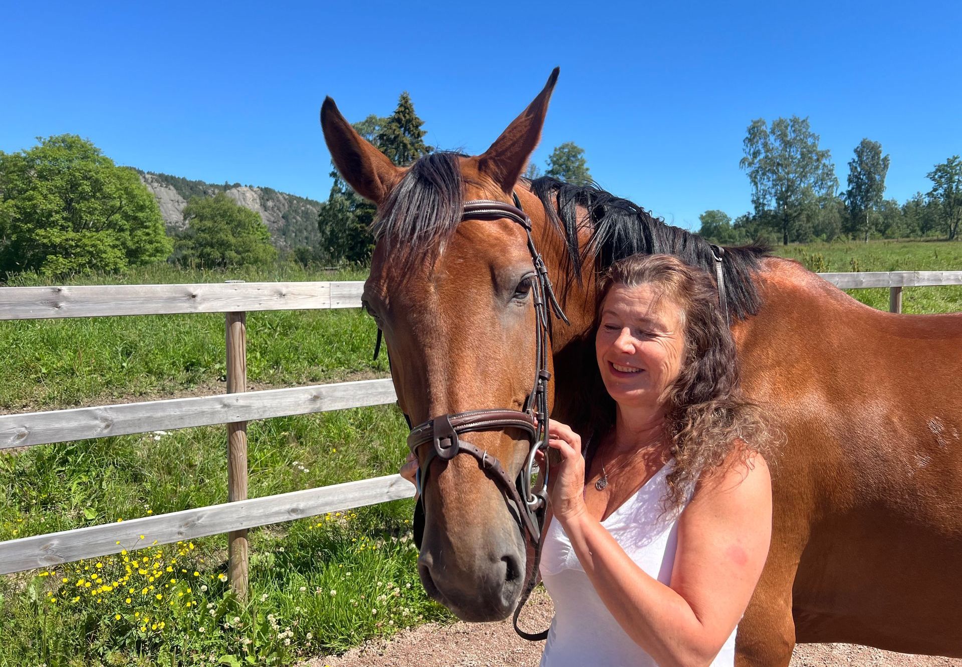 En kvinne står ved siden av en brun hest på et jorde.