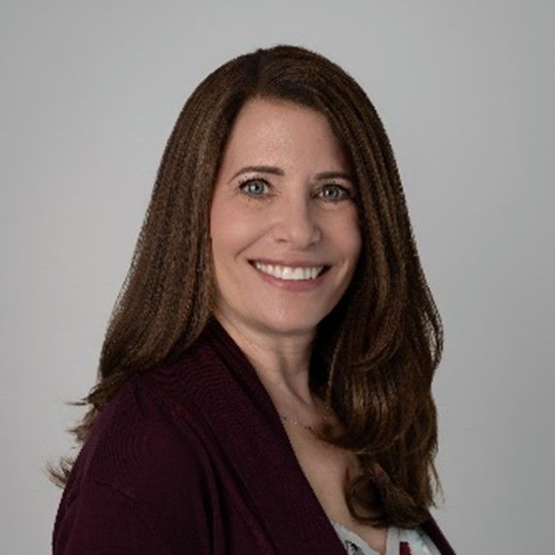 A woman with long brown hair is smiling and wearing a purple sweater.