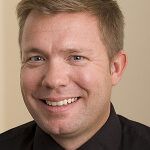 A man in a black shirt and tie is smiling for the camera.