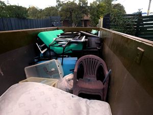 A dumpster filled with discarded items, including a mattress, plastic chairs, storage bins, and furniture.