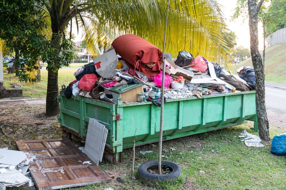 A large, overflowing green dumpster filled with household waste and debris, sitting in a grassy area near some trees.