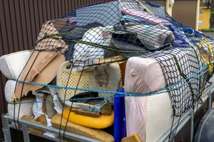 A utility trailer loaded with discarded mattresses, foam padding, and miscellaneous household items secured by a cargo net.
