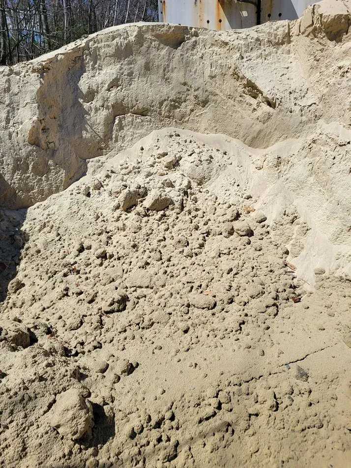 A large, sloped pile of light tan, coarse sand or sediment with visible clumps and uneven texture under bright sunlight.