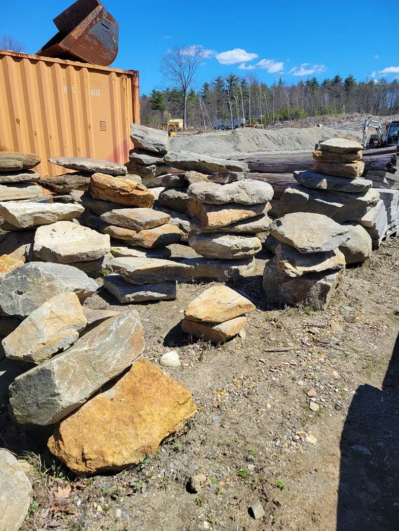 A yard filled with stacked flat stones and boulders next to a large orange shipping container under a clear blue sky.