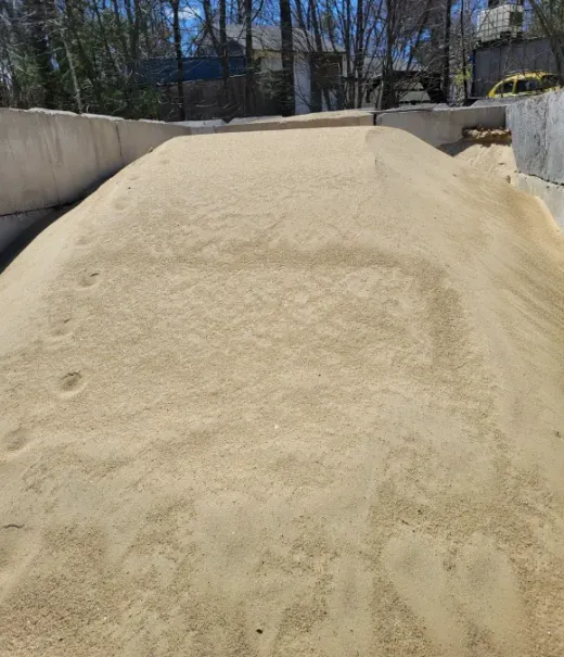 A large, tan-colored pile of sand sits between concrete walls under a sunny sky with trees in the background.