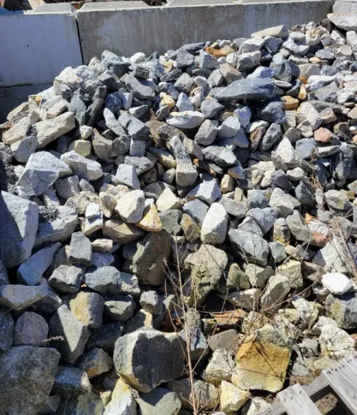 A large, high-angle pile of varied, light-to-dark grey rocks and stones outdoors in bright sunlight.