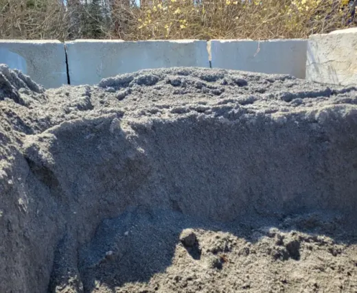 A large, gray mound of sand or soil sits in a bin enclosed by concrete barriers against a background of trees.