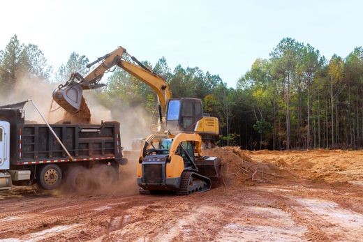 A yellow excavator loads red dirt into the back of a dump truck at a construction site near a forest.